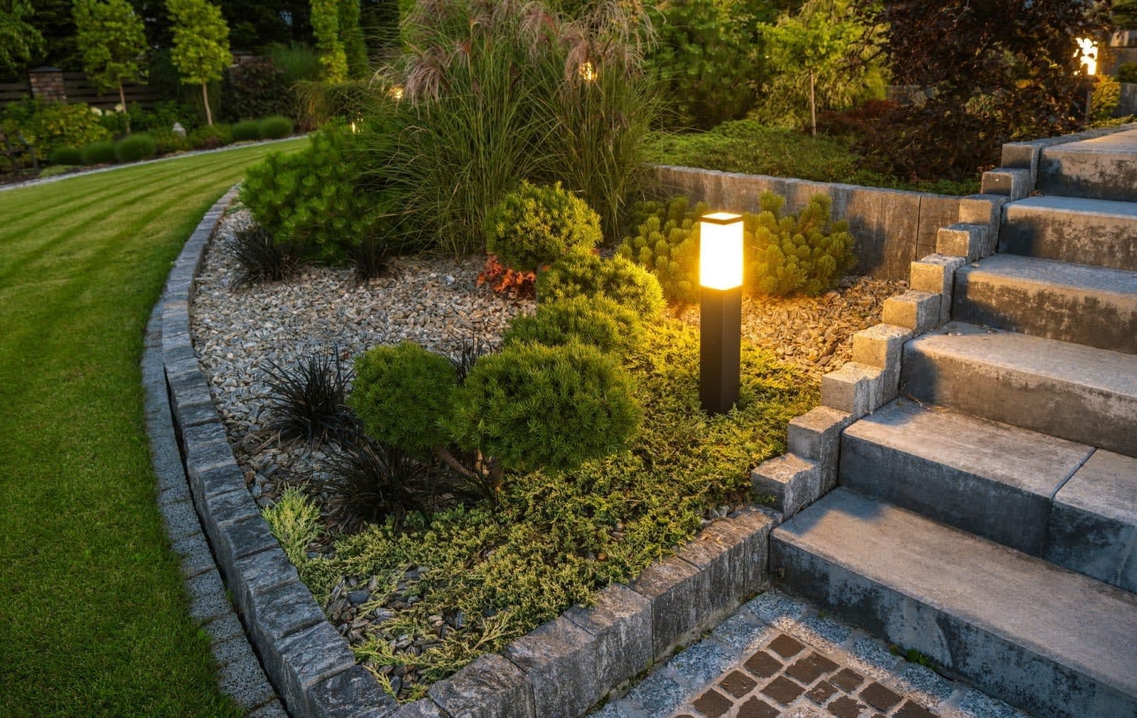 Brede natuurstenen trap in de avondschemering, naast een verhoogd grindbed met geschoren coniferen en een sokkellamp